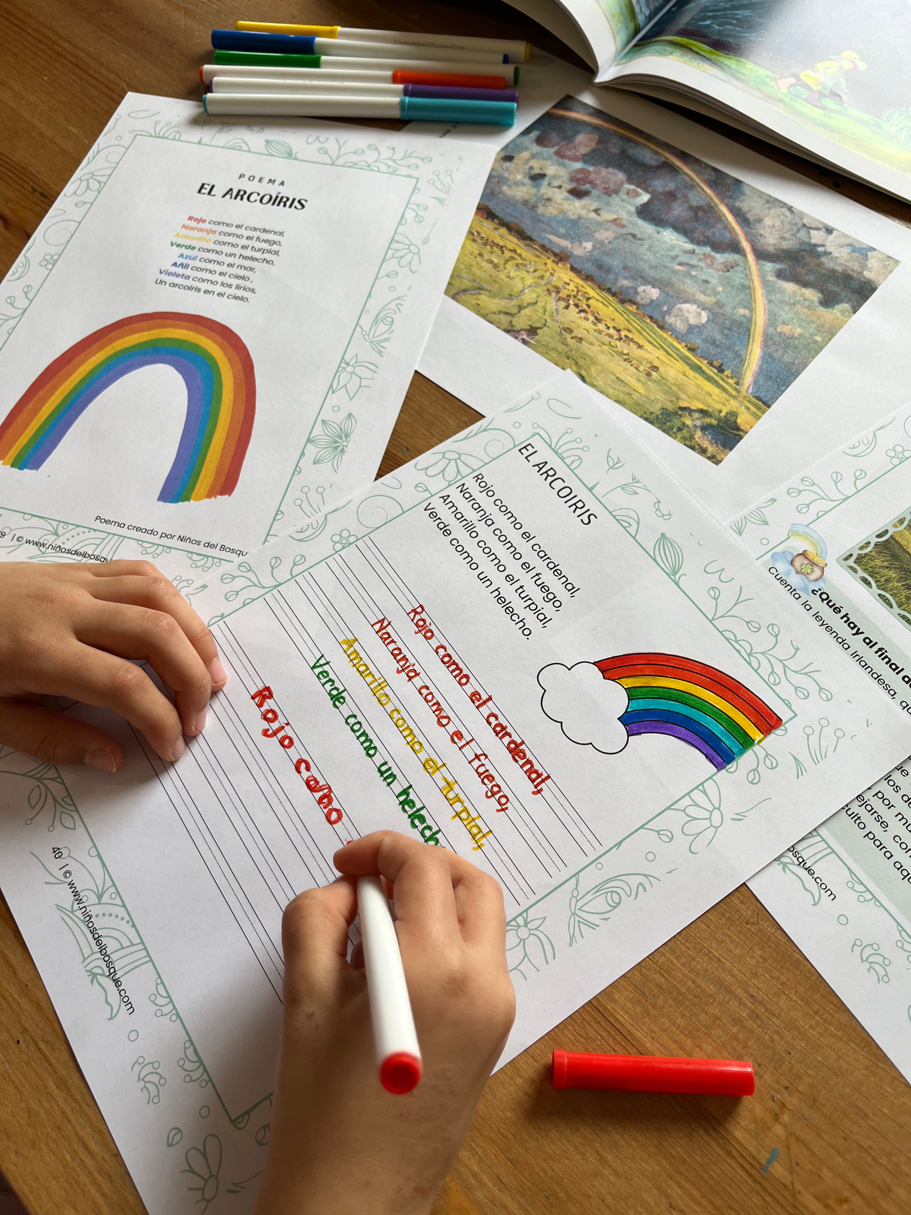 Niño escribiendo en una ficha educativa sobre el arcoíris con ilustraciones y actividades de naturaleza, acompañada de rotuladores de colores y una fotografía de un arcoíris