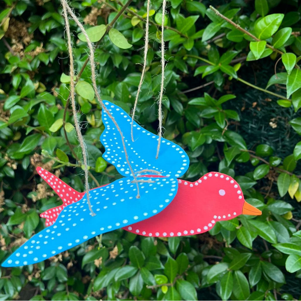 Manualidad infantil de un pájaro de colores colgante hecha con papel y cuerda, sostenida con la mano frente a un arbusto verde.